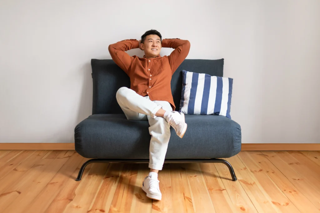 Smiling man sits with his arms behind his head on a couch after learning how to be a minimalist and save money